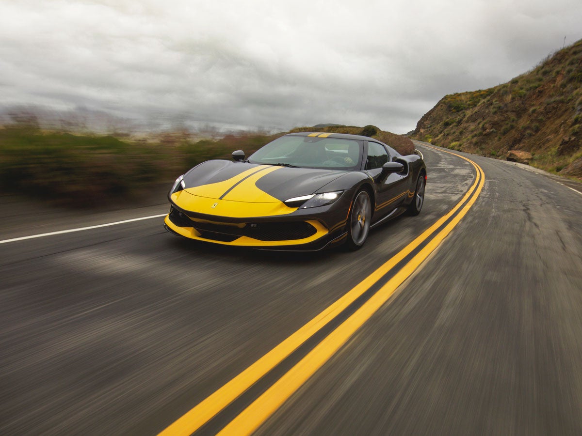 A black Ferrari with yellow stripes speeds along a coastal road under a cloudy sky.