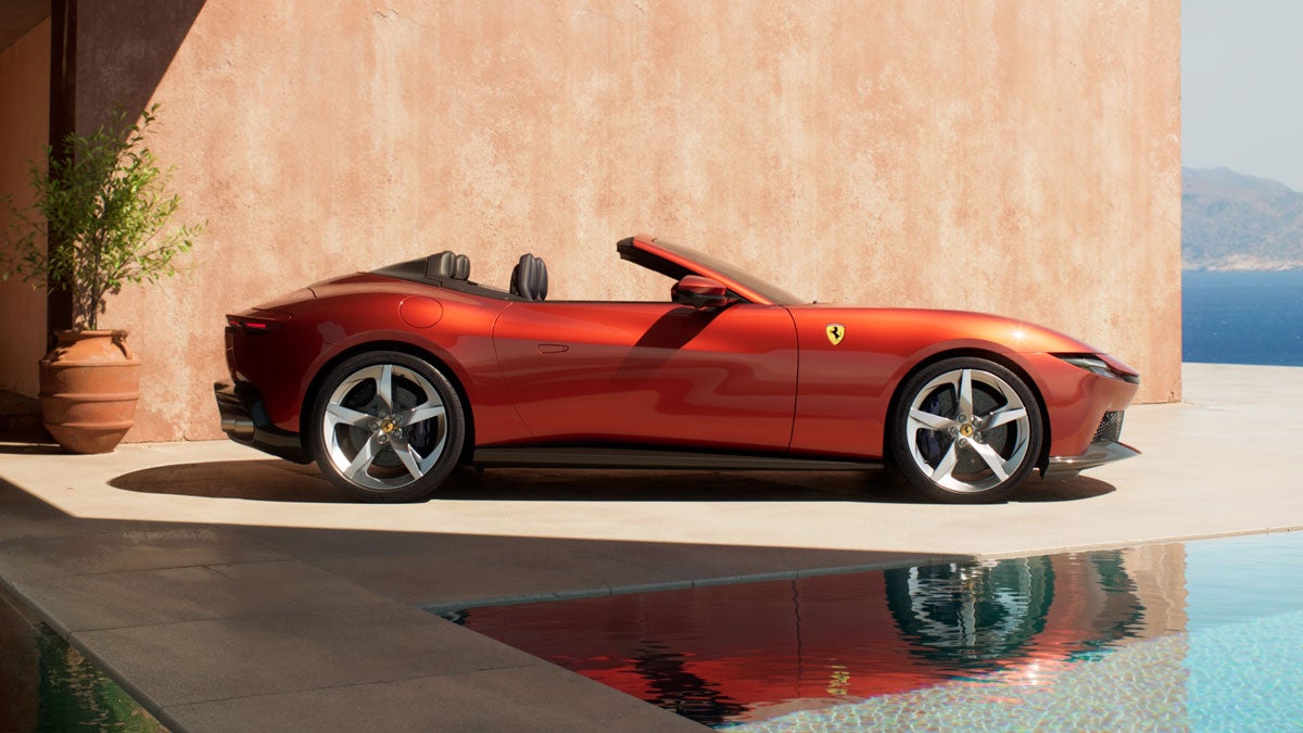 Sleek red Ferrari Roma Spider convertible parked beside a swimming pool with a terracotta wall and ocean in background.