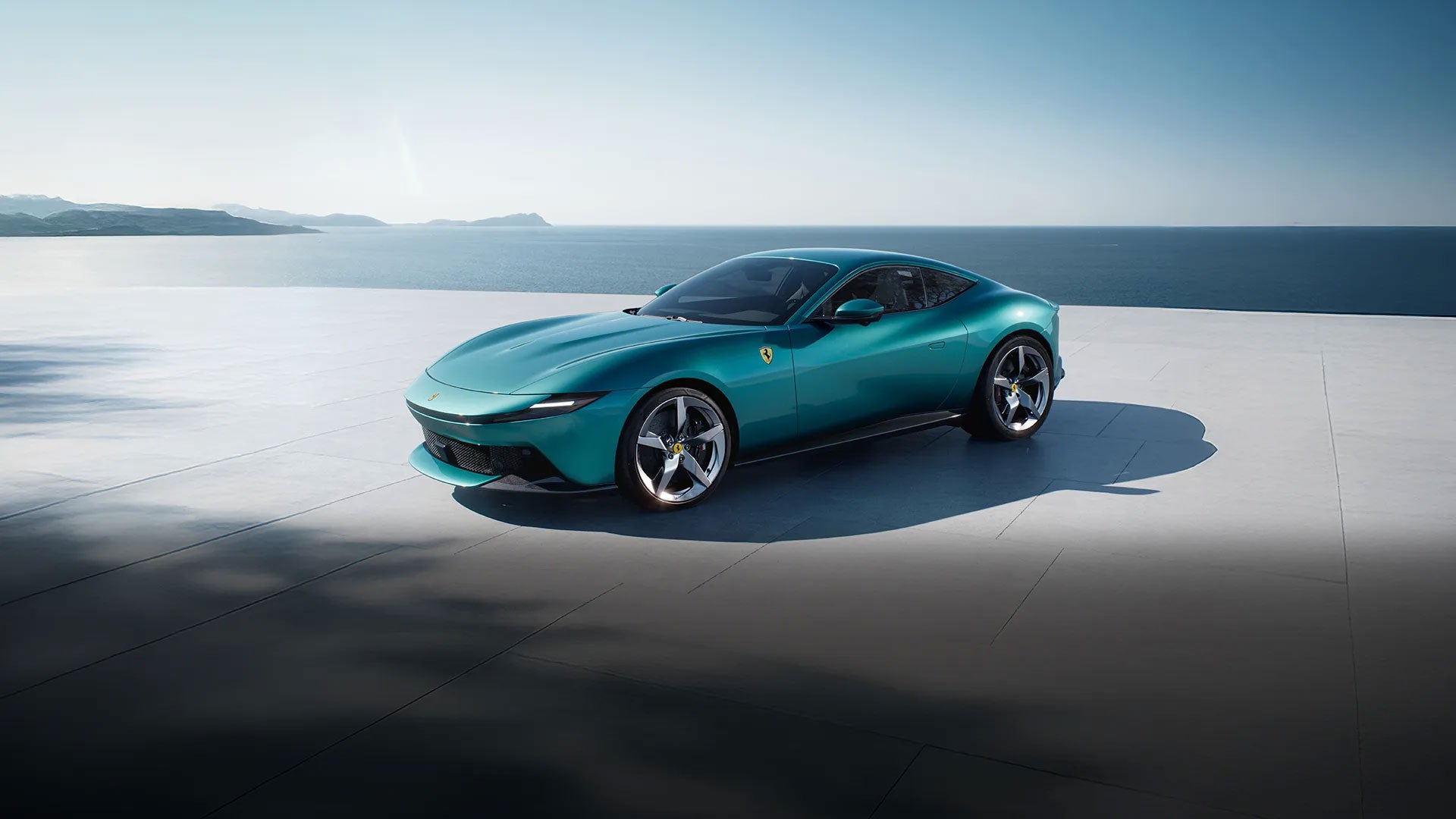 A dark turquoise Ferrari Roma on a light gray patio overlooking a calm blue sea under a clear sky.
