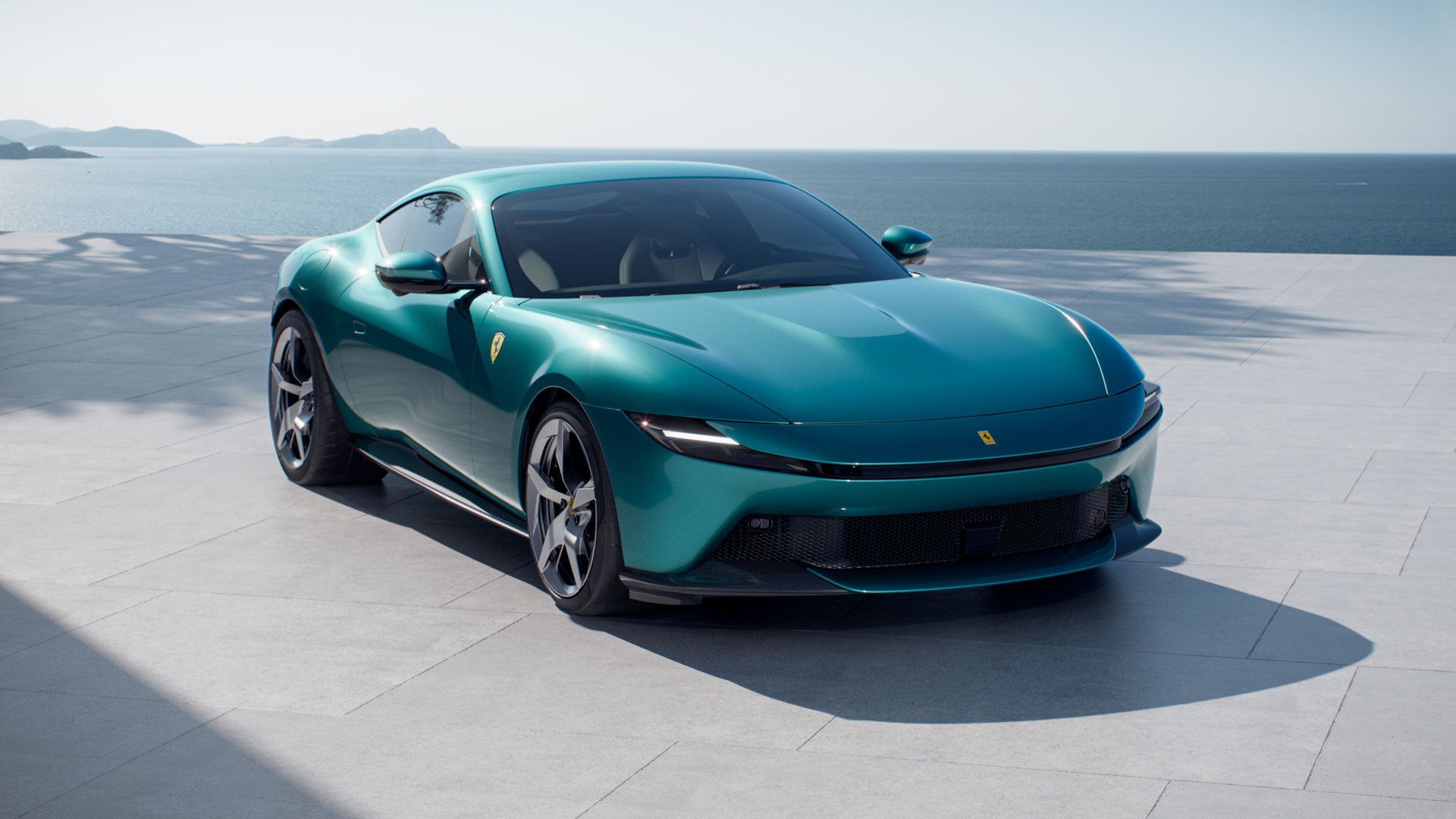 A teal Ferrari parked on a stone patio overlooking the ocean and mountains.