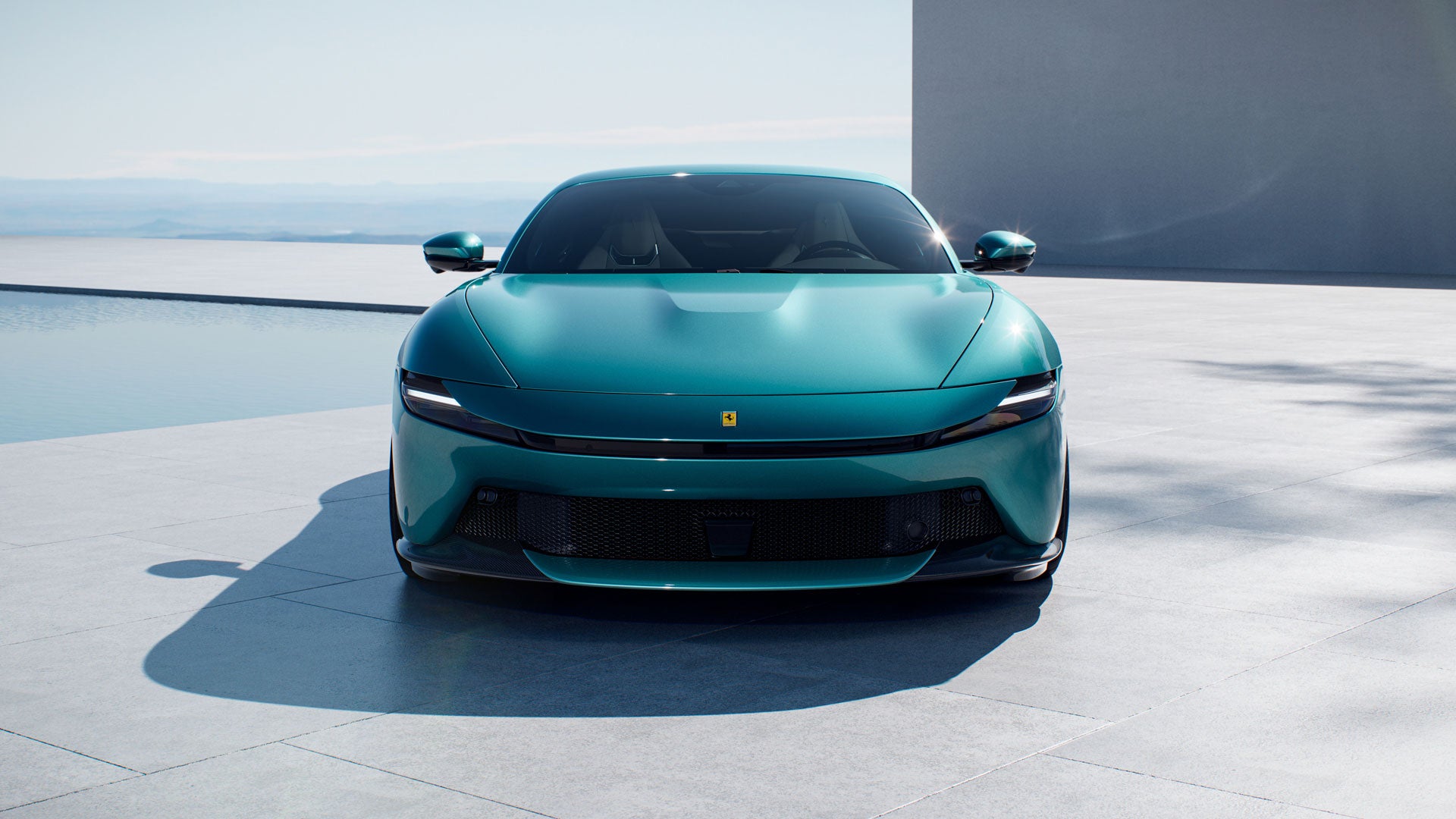 Front view of a teal green Ferrari parked on a light-colored patio next to a calm body of water.