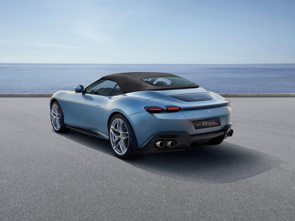 Pale blue convertible Ferrari Roma Spider with a black soft top parked on asphalt by the ocean.