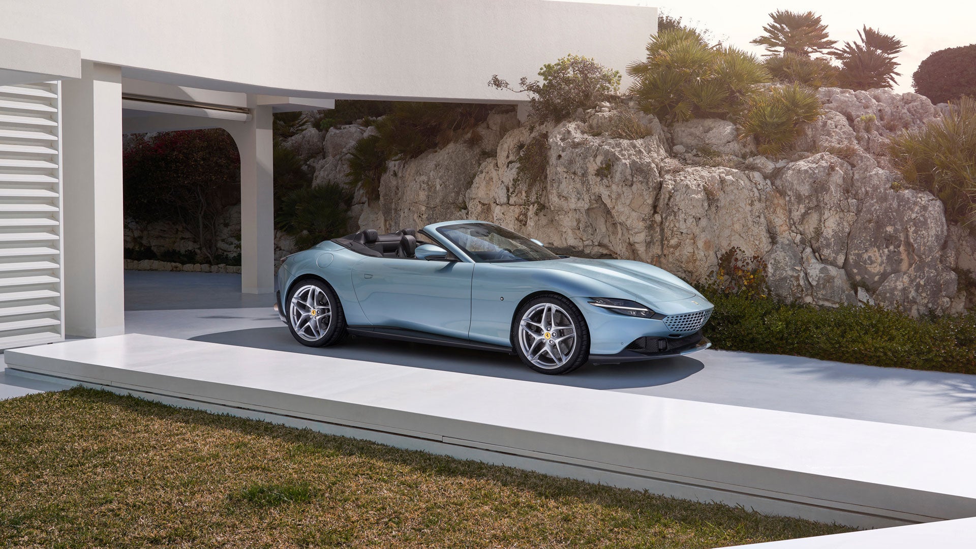 A light blue convertible car sits on a modern white driveway, partially shaded by an architectural structure, under a clear sky.